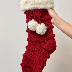 May include: A red crocheted Christmas stocking with a white faux fur cuff and two white pom-poms.