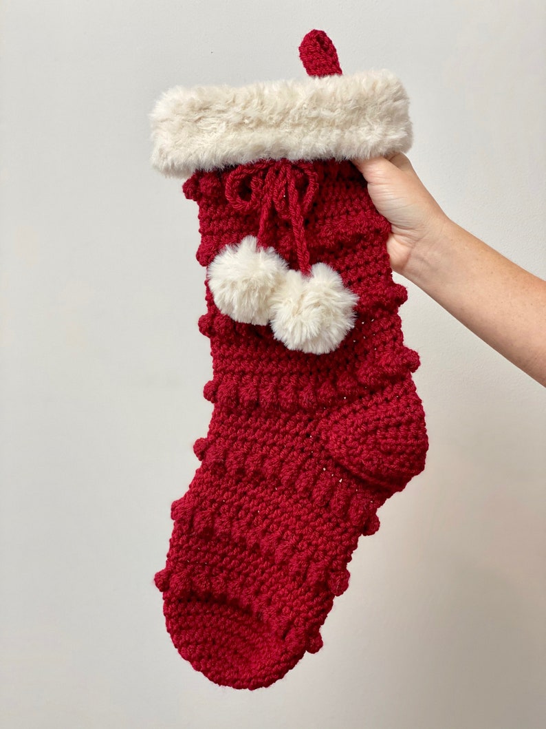 May include: A red crocheted Christmas stocking with a white faux fur cuff and two white pom-poms.