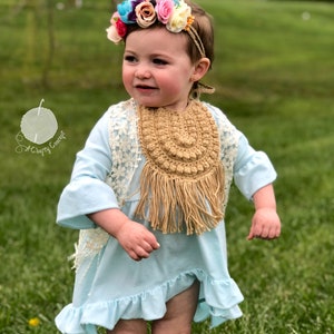 Caucasian baby girl wearing a dark tan crochet baby bib, a blue shirt and a flower crown.  Listing is for a crochet bib pattern and not the finished baby bib.