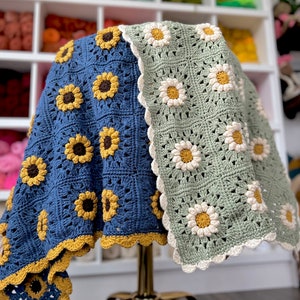 May include: A crocheted blanket with a blue and green background featuring a sunflower and daisy pattern. The blanket has a scalloped edge.