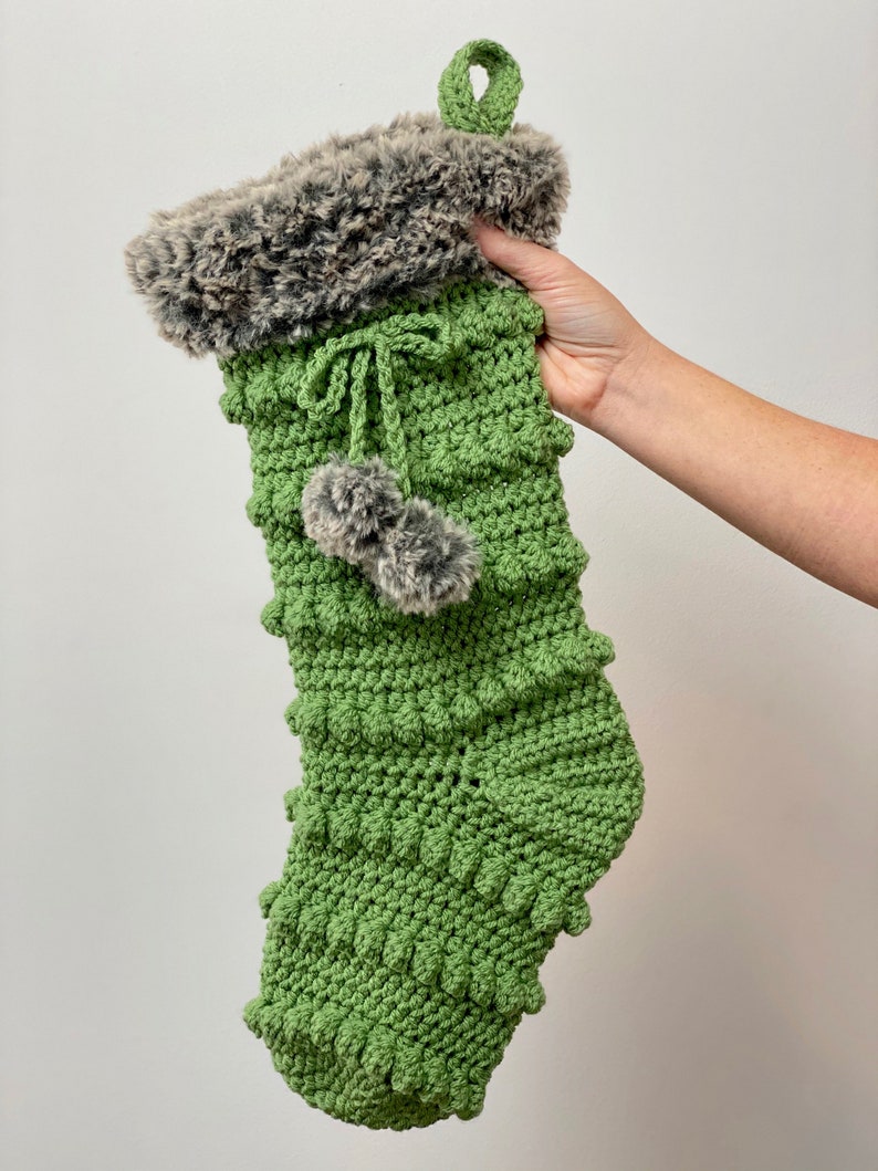 May include: Green crocheted Christmas stocking with a faux fur cuff and two gray pom-poms.