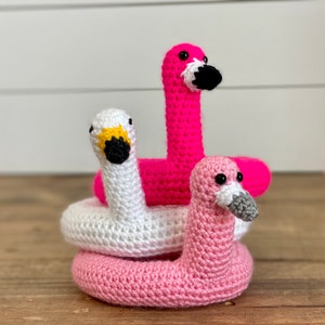 Op de afbeelding: Drie gehaakte flamingo zwembandjes, één roze, één wit en één kleinere roze. De zwembandjes zijn op elkaar gestapeld.