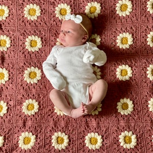 May include: A baby wearing a white onesie and a white bow headband lies on a pink crocheted blanket with a daisy pattern.
