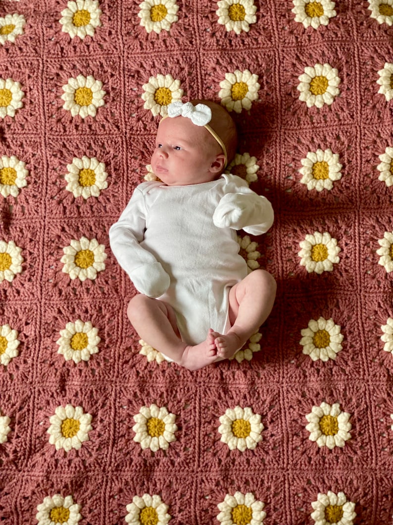 May include: A baby wearing a white onesie and a white bow headband lies on a pink crocheted blanket with a daisy pattern.