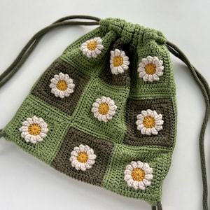 Könnte beinhalten: Eine grüne und braune gehäkelte Kordeltasche mit einem Gänseblümchenmuster. Die Tasche hat einen langen, verstellbaren Riemen.