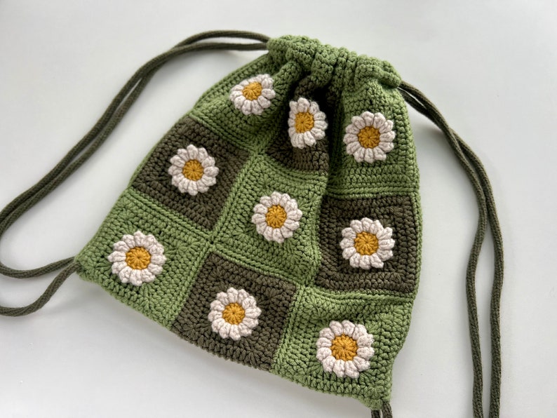 Puede incluir: Una bolsa de cord&oacute;n de crochet verde y marr&oacute;n con un patr&oacute;n de margarita. La bolsa tiene una correa larga y ajustable.