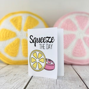 May include: A white card with the text "Squeeze the Day" in black lettering. The card features a yellow lemon slice and a pink grapefruit slice. The card is sitting on a white wooden surface with a yellow crocheted lemon and a pink crocheted grapefruit behind it.