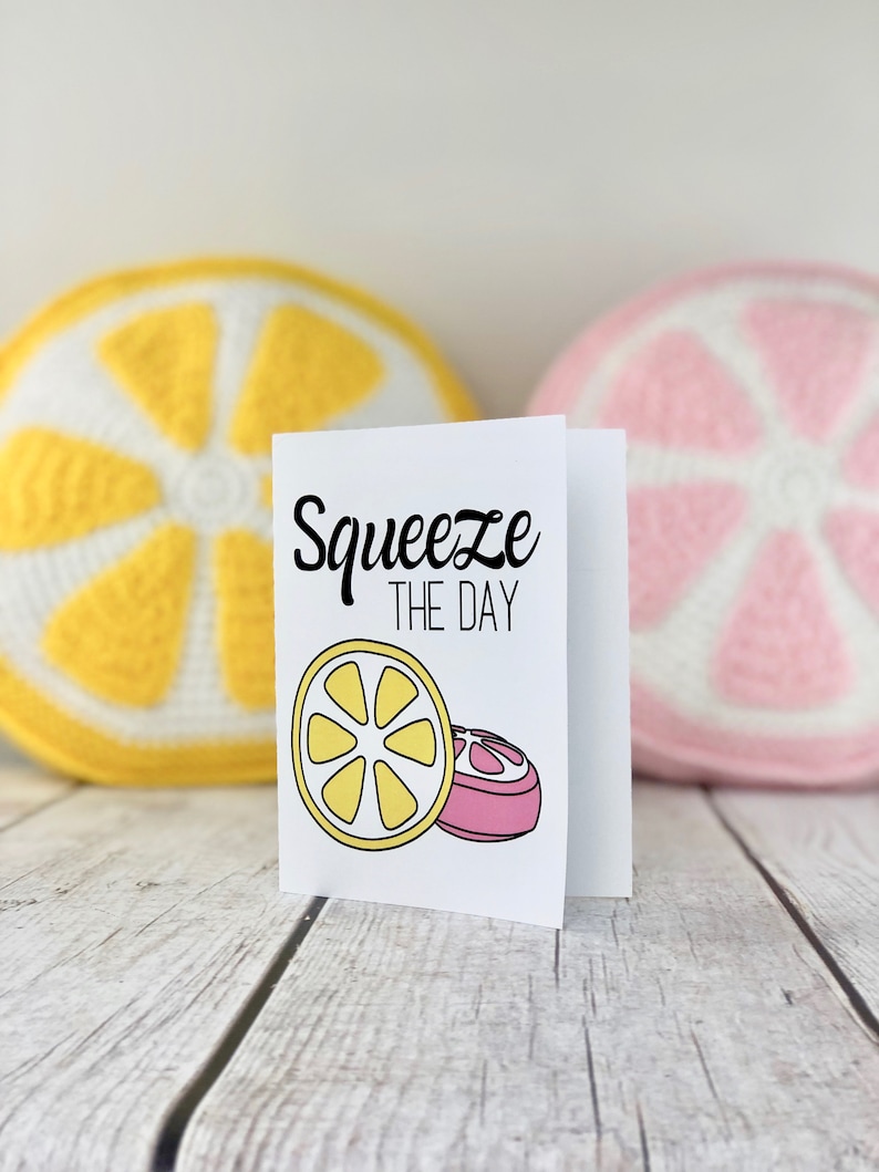 May include: A white card with the text "Squeeze the Day" in black lettering. The card features a yellow lemon slice and a pink grapefruit slice. The card is sitting on a white wooden surface with a yellow crocheted lemon and a pink crocheted grapefruit behind it.