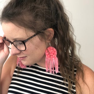 May include: A person wearing a black and white striped tank top with pink crocheted earrings. The earrings have a round, textured top and long fringe.