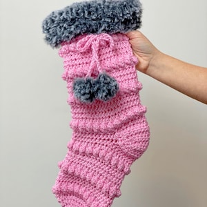 May include: Pink crocheted Christmas stocking with a gray faux fur cuff and pom poms.