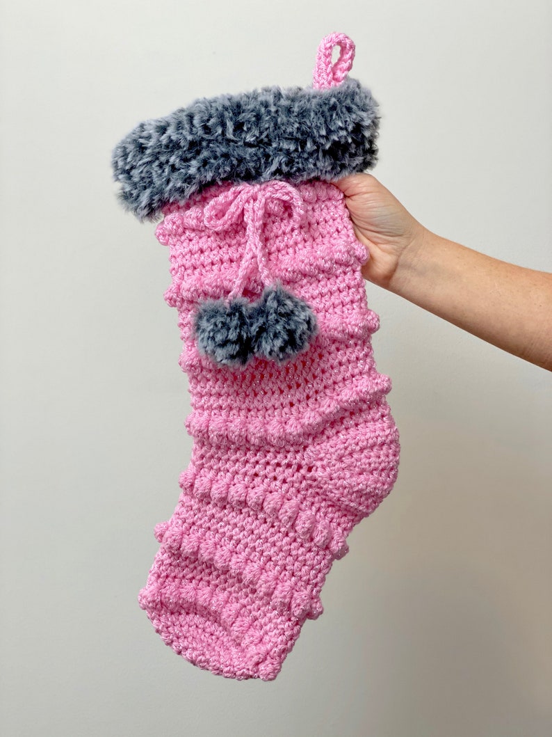 May include: Pink crocheted Christmas stocking with a gray faux fur cuff and pom poms.