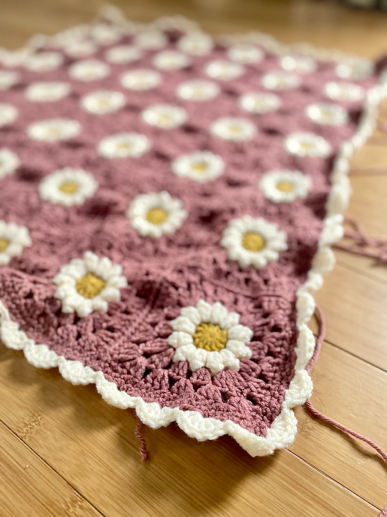 May include: A crocheted blanket with a pink background and white daisy flowers with yellow centers. The blanket has a white crocheted border.