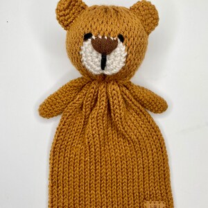 以下が含まれることがあります： 白い顔と茶色の鼻をした茶色のニットのクマ。クマは、"Wild Grace"と書かれた茶色の革タグが付いた茶色のニットの毛布に繋がっています。