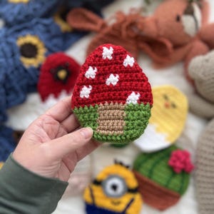 Peut inclure: Un champignon en crochet avec un chapeau rouge et des points blancs, une tige verte et une base brune. Le champignon est entouré d'autres objets en crochet, dont un minion jaune, un cactus et un poussin.