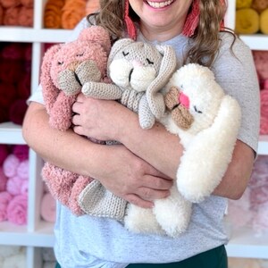 May include: Three handmade plush bunny rabbits in shades of pink, gray, and white. The bunnies have closed eyes, long ears, and textured fur. The bunnies are being held in front of a shelf filled with colorful yarn.