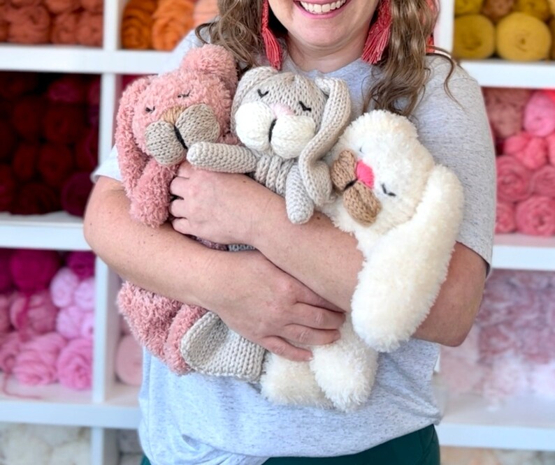 May include: Three handmade plush bunny rabbits in shades of pink, gray, and white. The bunnies have closed eyes, long ears, and textured fur. The bunnies are being held in front of a shelf filled with colorful yarn.