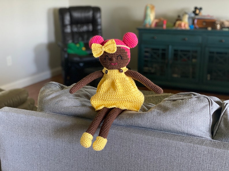 Crochet doll with dark skin wearing a yellow dress, yellow shoes and bright pink hair. Listing is for a crochet doll pattern and not the finished piece.