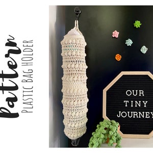 May include: A crocheted cream-colored plastic bag holder with a white plastic bag inside. The holder is hanging from a hook on a black wall. The wall also has a black letter board with the text "OUR TINY JOURNEY" in white letters.