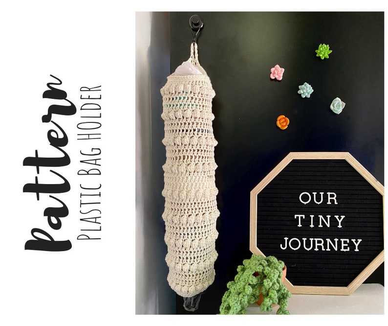 May include: A crocheted cream-colored plastic bag holder with a white plastic bag inside. The holder is hanging from a hook on a black wall. The wall also has a black letter board with the text "OUR TINY JOURNEY" in white letters.