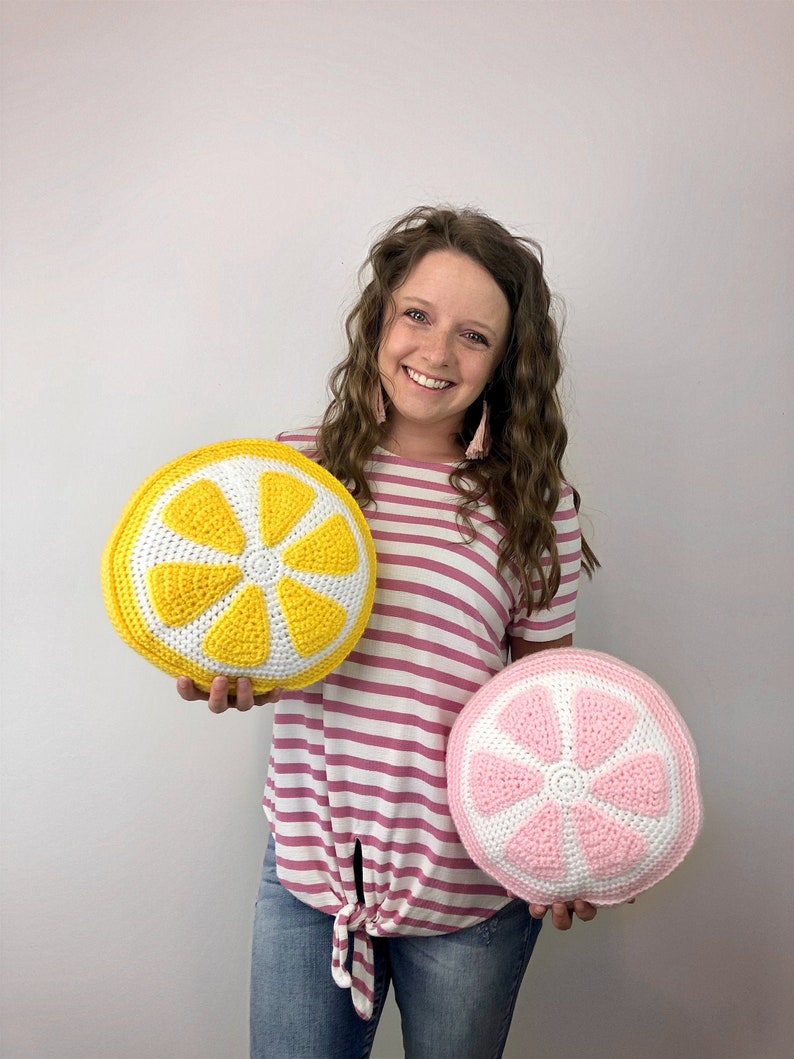 May include: Two crocheted lemon slice pillows, one yellow and one pink, held by a person wearing a pink and white striped shirt and blue jeans.