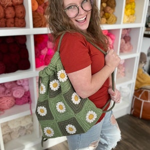 Może przedstawiać: Zielona torba ze sznurkiem do ściągania wykonana na szydełku z białymi kwiatami stokrotek. Torbę trzyma osoba ubrana w czerwoną koszulę i niebieskie dżinsy.