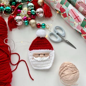 May include: A crocheted Santa Claus ornament with a red hat, white beard, and black eyes. The ornament is surrounded by yarn, wrapping paper, and Christmas decorations.