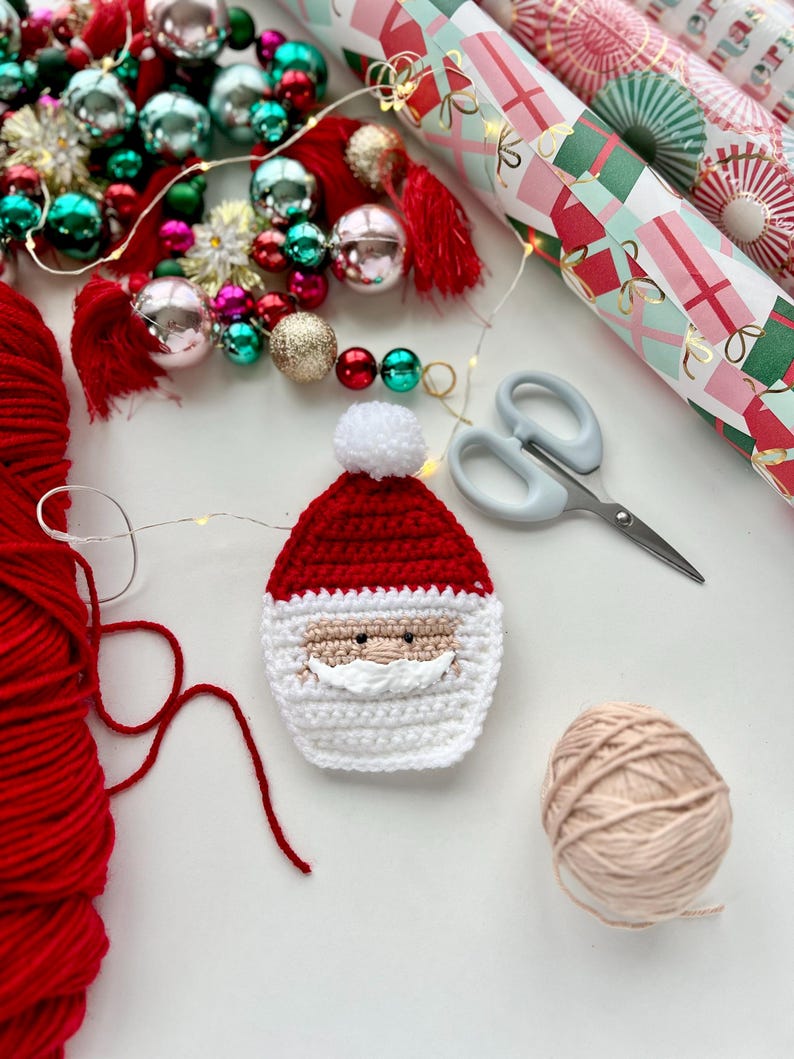 May include: A crocheted Santa Claus ornament with a red hat, white beard, and black eyes. The ornament is surrounded by yarn, wrapping paper, and Christmas decorations.