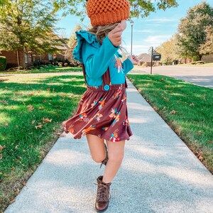 Op de afbeelding: Een jong persoon draagt een bruine en oranje pompoenmuts, een blauw shirt met lange mouwen met een bloemenprint, een bruine rok met bloemenprint en bruine laarzen. De persoon loopt op een stoep.