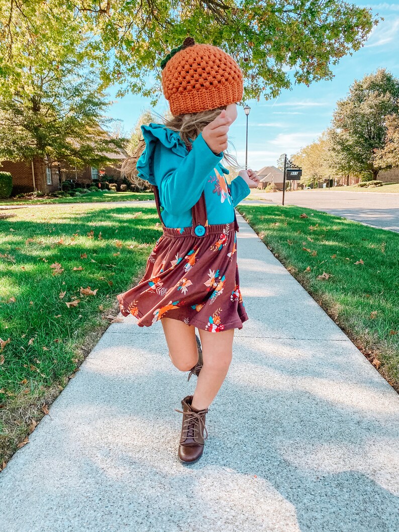 Op de afbeelding: Een jong persoon draagt een bruine en oranje pompoenmuts, een blauw shirt met lange mouwen met een bloemenprint, een bruine rok met bloemenprint en bruine laarzen. De persoon loopt op een stoep.