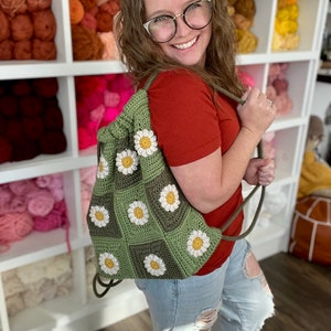 Puede incluir: Una bolsa de cord&oacute;n verde hecha a crochet con flores de margarita blancas. La bolsa la lleva una persona que lleva una camisa roja y unos vaqueros azules.