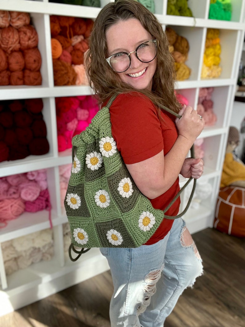 Puede incluir: Una bolsa de cord&oacute;n verde hecha a crochet con flores de margarita blancas. La bolsa la lleva una persona que lleva una camisa roja y unos vaqueros azules.