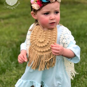 Caucasian baby girl wearing a dark tan crochet baby bib, a blue shirt and a flower crown.  Listing is for a crochet bib pattern and not the finished baby bib.