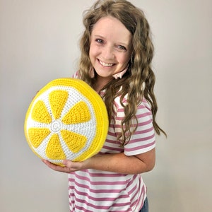 May include: A woman is holding a crocheted lemon-shaped pillow. The pillow is yellow with a white center and is made with a textured yarn.