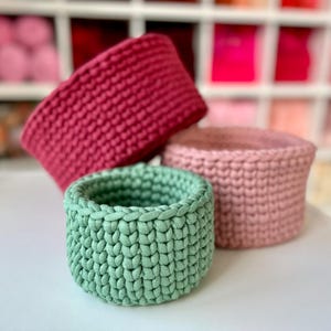May include: Three handmade crochet baskets in varying shades of pink and green. The front basket is a light green, the middle basket is a soft pink, and the back basket is a deep rose color. The baskets are cylindrical and have a textured appearance.