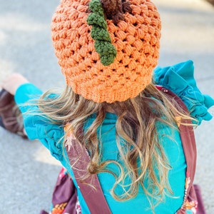 Op de afbeelding: Een jong persoon draagt een oranje-bruine gebreide pompoenmuts met groene en bruine accenten. De muts wordt gedragen over lang blond haar. De persoon draagt een turquoise shirt en een bruine bloemenjurk met bruine banden.