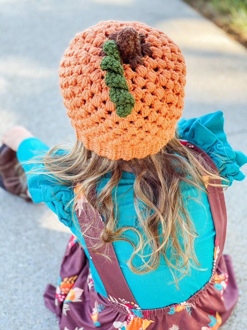 Op de afbeelding: Een jong persoon draagt een oranje-bruine gebreide pompoenmuts met groene en bruine accenten. De muts wordt gedragen over lang blond haar. De persoon draagt een turquoise shirt en een bruine bloemenjurk met bruine banden.