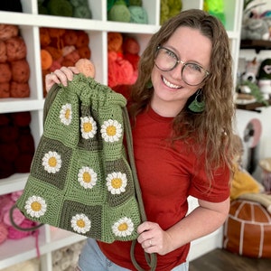 Puede incluir: Una bolsa de cord&oacute;n verde hecha a crochet con flores de margarita blancas. La bolsa la sostiene una persona que lleva una camisa roja y pantalones vaqueros azules.