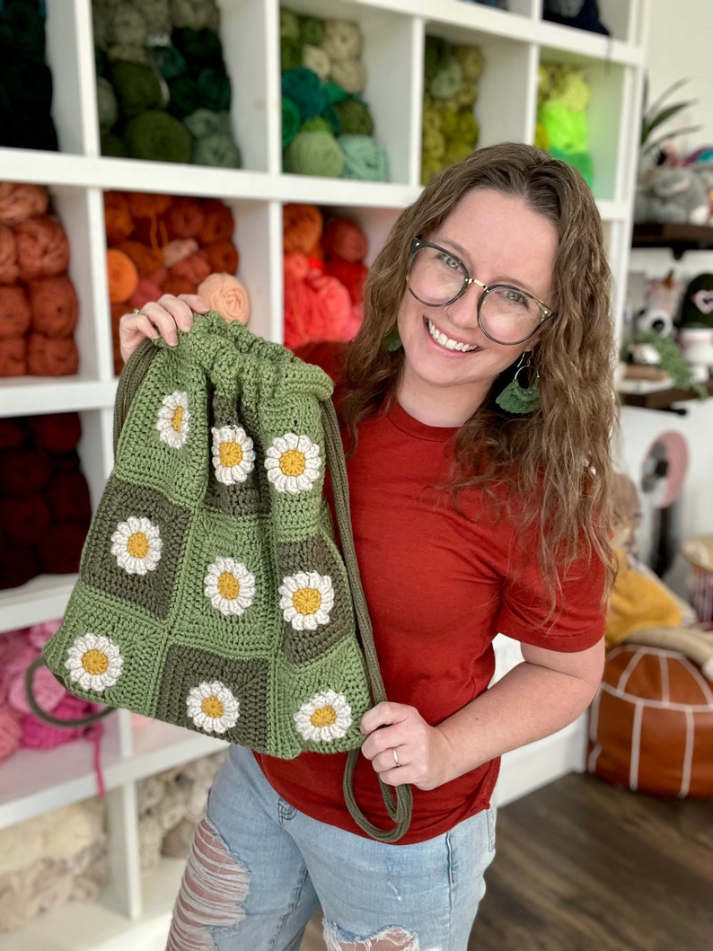 Puede incluir: Una bolsa de cord&oacute;n verde hecha a crochet con flores de margarita blancas. La bolsa la sostiene una persona que lleva una camisa roja y pantalones vaqueros azules.