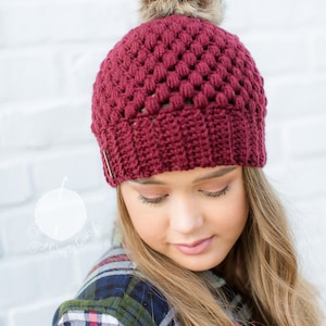 crochet beanie modeled by beautiful blond woman in the color maroon with a brown faux fur pom pom on top