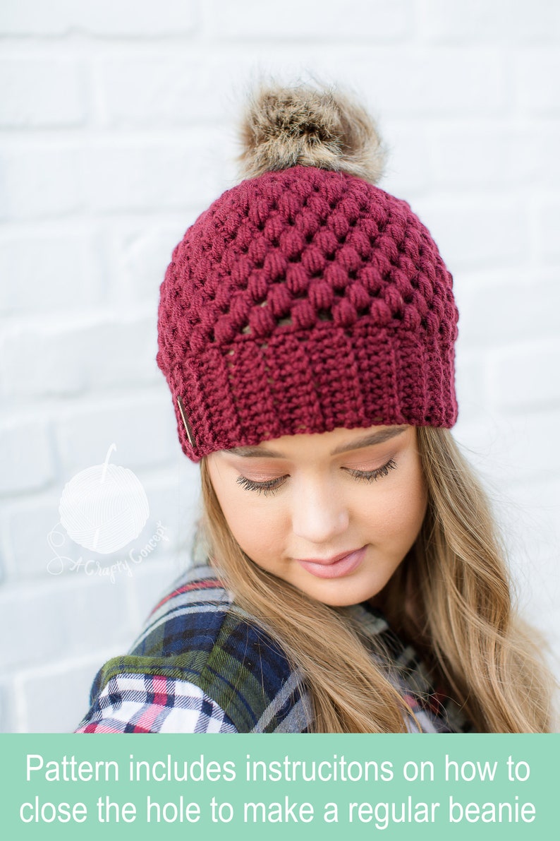 crochet beanie modeled by beautiful blond woman in the color maroon with a brown faux fur pom pom on top