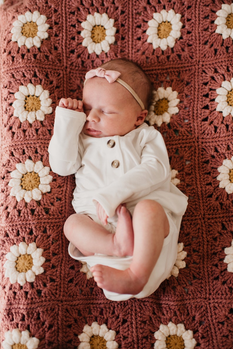 Baby laying on daisy granny square blanket with a pink background, white petals, and a yellow center. The listing is for a digital download crochet pattern and not the finished blanket.
