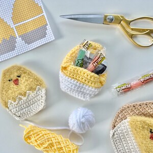 K&ouml;nnte beinhalten: Ein Bastelprojekt mit geh&auml;kelten K&uuml;ken- und Ei-Formen, einem geh&auml;kelten Korb gef&uuml;llt mit S&uuml;&szlig;igkeiten und einem H&auml;kelmuster auf einem Blatt Papier. Die S&uuml;&szlig;igkeiten sind Sour Punch-Strohhalme.