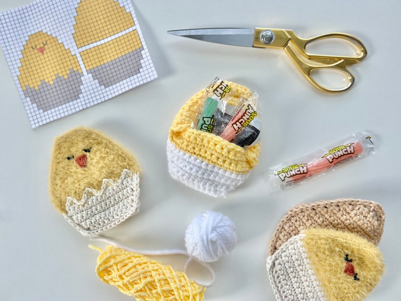 K&ouml;nnte beinhalten: Ein Bastelprojekt mit geh&auml;kelten K&uuml;ken- und Ei-Formen, einem geh&auml;kelten Korb gef&uuml;llt mit S&uuml;&szlig;igkeiten und einem H&auml;kelmuster auf einem Blatt Papier. Die S&uuml;&szlig;igkeiten sind Sour Punch-Strohhalme.