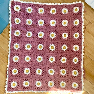 May include: A crocheted blanket with a daisy pattern in white and yellow on a mauve background. The blanket has a white crocheted border.