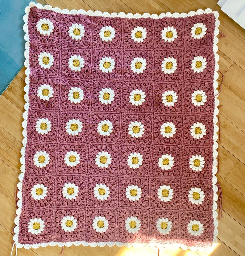 May include: A crocheted blanket with a daisy pattern in white and yellow on a mauve background. The blanket has a white crocheted border.