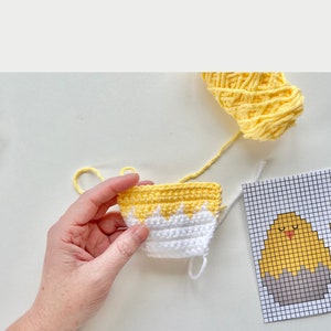 K&ouml;nnte beinhalten: Nahaufnahme eines geh&auml;kelten gelben und wei&szlig;en K&uuml;ken mit einem wei&szlig;en Garn-Schwanz. Das K&uuml;ken wird von einer Hand gehalten. Ein Graphenpapier-Muster f&uuml;r ein geh&auml;keltes K&uuml;ken ist im Hintergrund sichtbar.