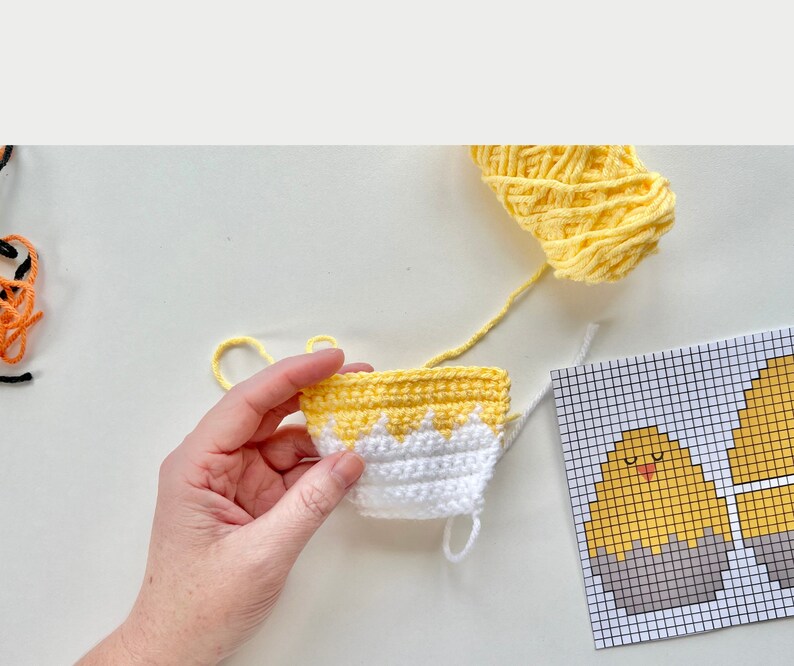 K&ouml;nnte beinhalten: Nahaufnahme eines geh&auml;kelten gelben und wei&szlig;en K&uuml;ken mit einem wei&szlig;en Garn-Schwanz. Das K&uuml;ken wird von einer Hand gehalten. Ein Graphenpapier-Muster f&uuml;r ein geh&auml;keltes K&uuml;ken ist im Hintergrund sichtbar.