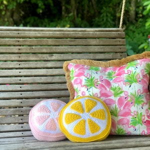 May include: Two crocheted lemon-shaped pillows on a wooden bench. One pillow is yellow and white, the other is pink and white. The pillows are sitting on a wooden bench with a pink flamingo patterned pillow behind them.