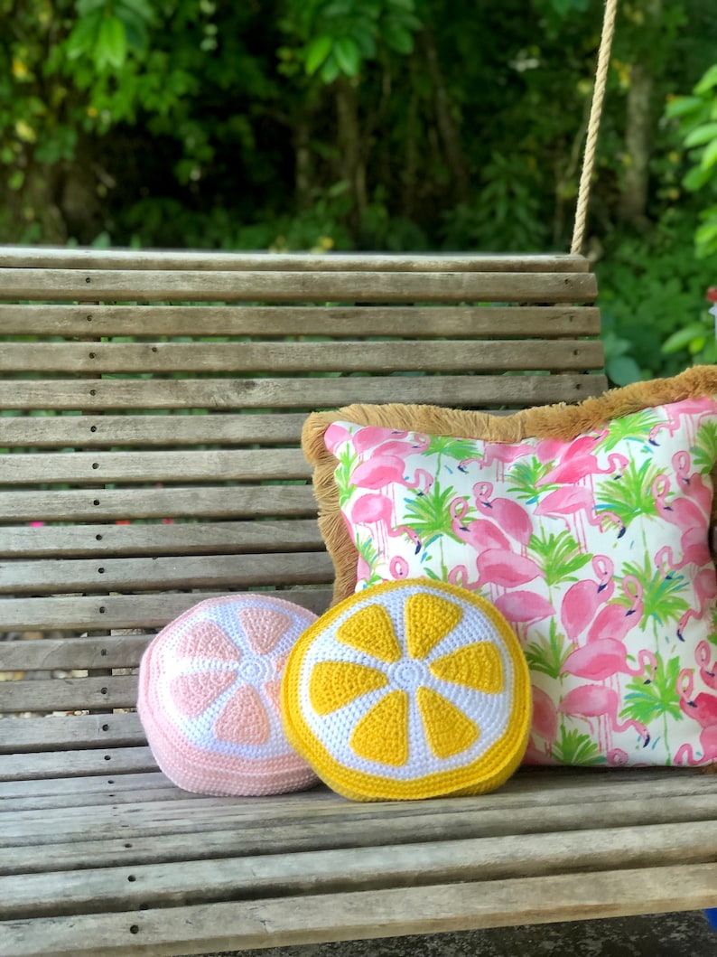 May include: Two crocheted lemon-shaped pillows on a wooden bench. One pillow is yellow and white, the other is pink and white. The pillows are sitting on a wooden bench with a pink flamingo patterned pillow behind them.