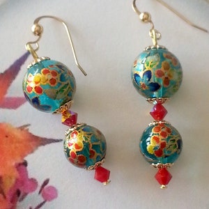 Op de afbeelding: Hangende oorbellen met turquoise bloemvormige kralen en gouden accenten. Rode kristallen kralen en gouden haken maken het ontwerp compleet. De tekst "Brigitte Créations" is zichtbaar.